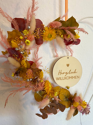 Weißer Herzhoop mit Trockenblumen in pink, gelb und rosa Tönen gebunden und Holzschild mit der Aufschrift Herzlich Willkommen in der Mitte hängend.