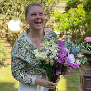 Mindful Movement Portrait von Marleen Jansen im Garten stehend, mit einem Blumenstrauß in der Hand und in die Kamera lachend. Sie trägt passend farbige Kleidung zum Gartenhintergrund. Die Sonne scheint und es ist ein Sommertag.