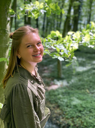 Mindful Movement Portrait von Marleen Jansen im Wald stehend und in die Kamera lachend. Sie trägt passend farbige Kleidung zum Waldhintergrund. Die Sonne scheint und es ist ein Sommertag.