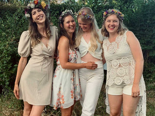 Jungesell:innen Abschied - Teameevent - 4 hellgekleidete Frauen mit Blumenschmuck auf dem Kopf, die in die Kamera lachen, sich freuen und vor einer grünen Hecke stehen.