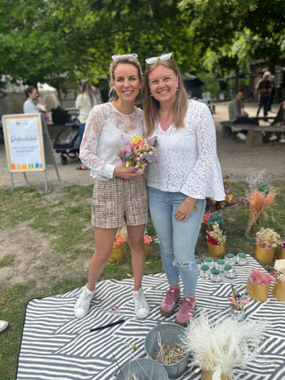 Mumevents Gruenderin und Marleen Jansen von Mindful Movement stehen zusammn beim Picknick und lächeln in die Kamera. Eine trägt eine Discokugel in der Hand mit Trockenblumen dekoriert.