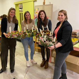 4 Frauen halte ihre Adventskränze mit Kerzen in der Hand und lächeln in die Kamera beim Adventskranz Workshop in Münster.