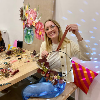 Workshop Teilnehmerin mit eigenem gebasteltem Blumenhoop in Hand. Die Frau mitte 20, mit blonden Haaren, Brille, Jeanshose und hellem Pullover lächelt in die Kamera. Sie sitzt auf einer Holzbank an einem Holztisch auf dem viele Blumenreste, Faden, Schere und Band vom Basteln liegen. Im Hintergrund steht ein Gemälde auf Leinwand von Blumen in einem Glas. An dem Bild hängt ebenfalls ein Hoop. Hinter der Frau liegt ein gestreiftes Kissen. 