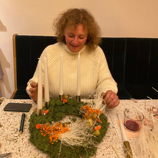 Eine lachende Frau hält ihren eigenen gestalteten Adventskranz mit Trockenblumen, Moos und Kerzen in einem DIY Workshop in die Kamera. Diese sind Beispielexemplare für den Blumen-Adventskranz-Workshop mit Trockenblumen in Münster.