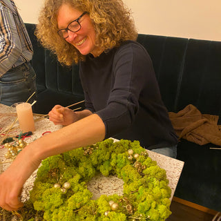 Eine lachende Frau hält ihren eigenen gestalteten Adventskranz mit Trockenblumen, Moos und Kerzen in einem DIY Workshop in die Kamera. Diese sind Beispielexemplare für den Blumen-Adventskranz-Workshop mit Trockenblumen in Münster.