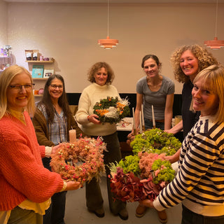 sechs lachende Frauen halten ihre eigenen gestalteten Adventskränze mit Trockenblumen, Moos und Kerzen in einem DIY Workshop in die Kamera. Diese sind Beispielexemplare für den Blumen-Adventskranz-Workshop mit Trockenblumen in Münster.