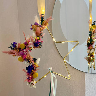 im Blumenworkshop gebastelter Sternhoop zum Aufhängen - DIY
Der große Sternenförmige Anhänger aus Metall, hat seitlich ein farbenfrohes Blumenarrangement dran gebunden. Zudem ist er umwickelt von einer Lichterkette. 