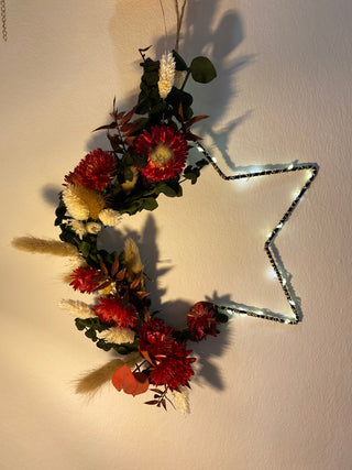 im Blumenworkshop gebastelter Sternhoop zum Aufhängen - DIY
Der große Sternenförmige Anhänger aus Metall, hat seitlich ein rot, grünes Blumenarrangement dran gebunden. Zudem ist er umwickelt von einer Lichterkette. 