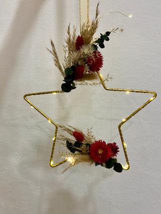 im Blumenworkshop gebastelter Sternhoop zum Aufhängen - DIY
Der große Sternenförmige Anhänger aus Metall, hat seitlich ein rot, beige, grünes Blumenarrangement dran gebunden und hängt an einem weiß-goldenen Schleifenband. Zudem ist er umwickelt von einer Lichterkette. 