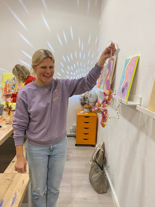 Eine lachende Frau hält ihren eigenen gestalteten Sternhoop aus Metall mit farbenfrohen Trockenblumen in einem DIY Workshop in die Kamera. Diese sind Beispielexemplare für den weihnachtliche Sternhoop-Workshop mit Trockenblumen in Münster.