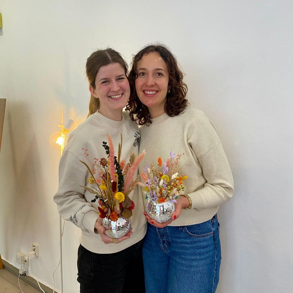 Zwei lachende Frauen halten ihre eigenen gestalteten Discokugeln mit Trockenblumen in einem DIY Workshop in die Kamera. Diese sind Beispielexemplare für den Blumen-Workshop mit Trockenblumen in Münster 