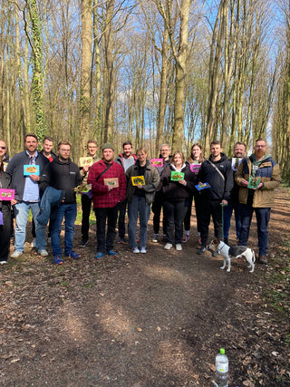 Ein Team beim Waldbaden in Münster mit Hund. Alle schauen in die Kamera und halten ihre Waldpostkarte in der Hand.